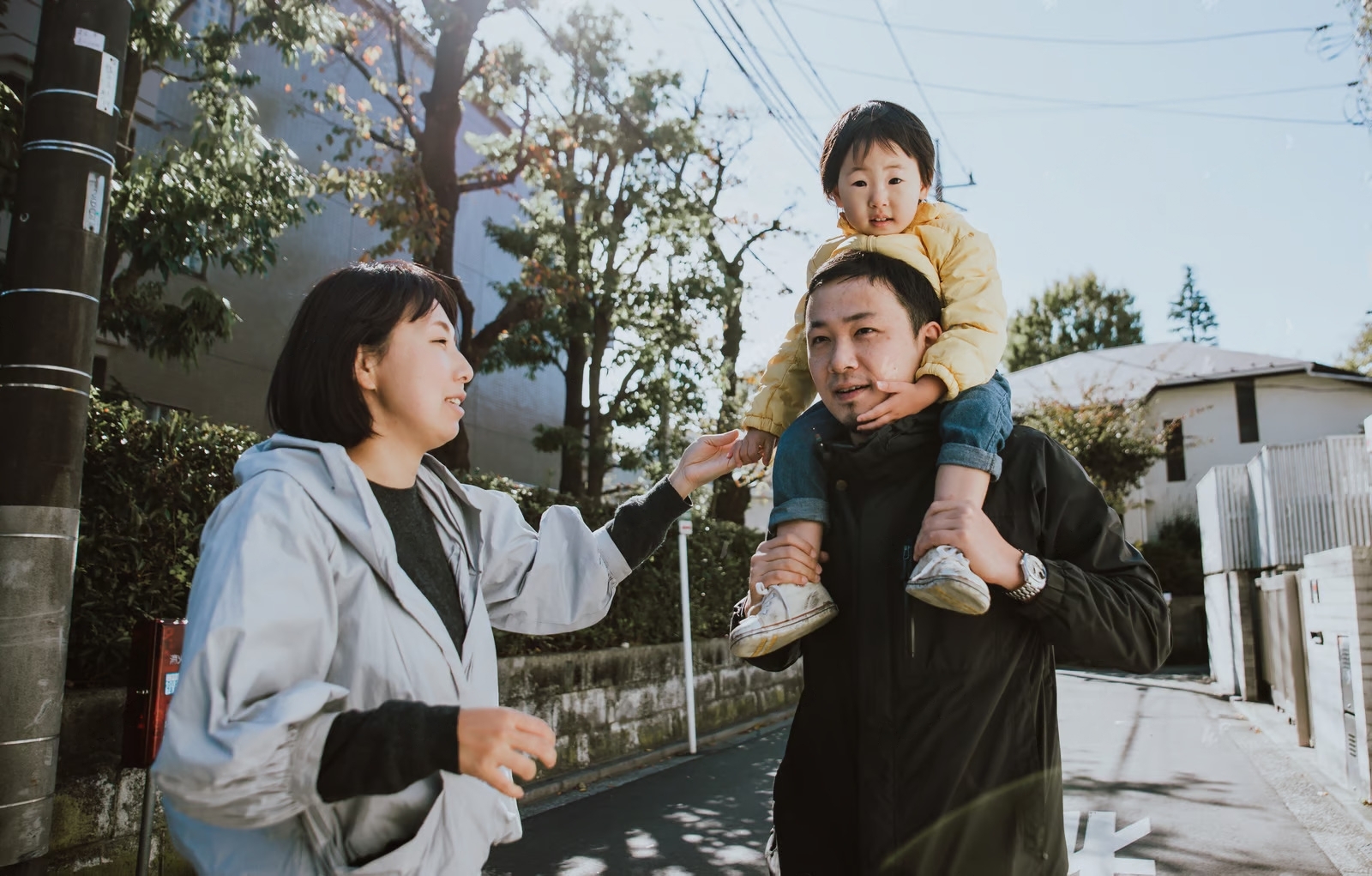 A man carries a child on his shoulders while walking on a sunny street. A woman walks beside them, reaching out towards the child. Trees and a building are in the background.