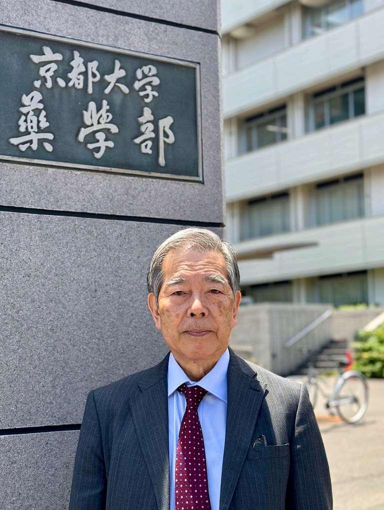 Our CEO in a suit stands outside next to a building wall bearing Japanese signage. The background shows part of a building with windows and a bicycle parked near stairs. The scene is in daylight.
